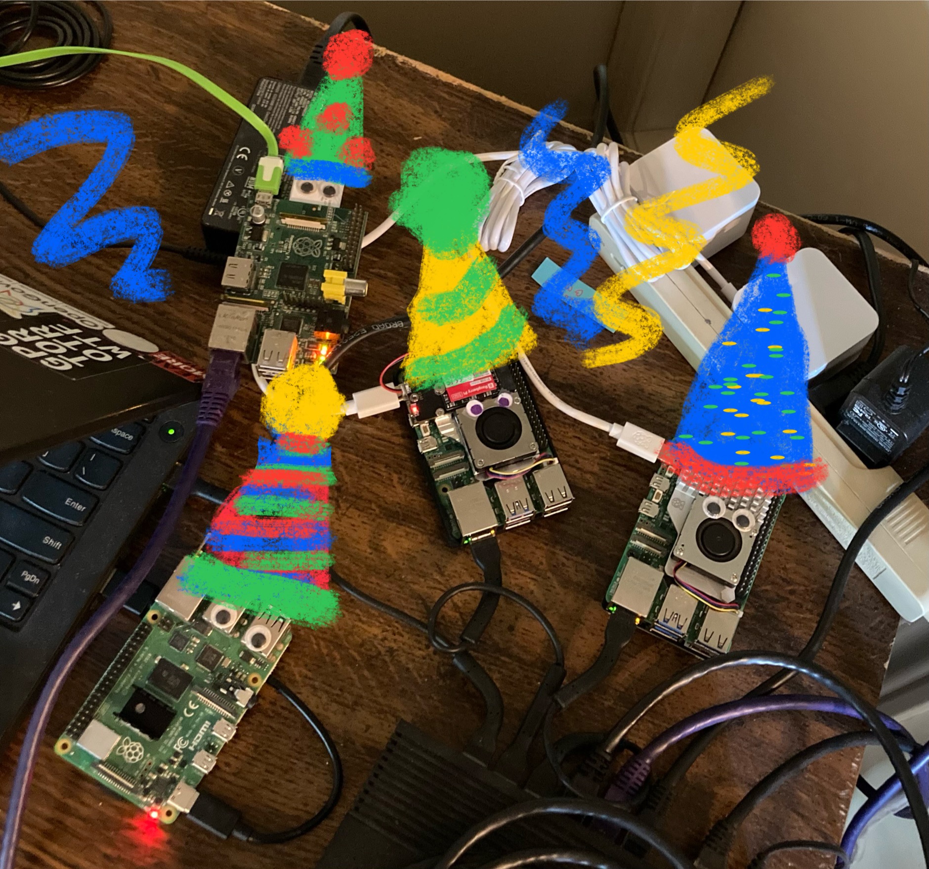 half-birthday portrait: four raspberry pi's with googly eyes on a wooden surface with messy networking and power cables around them. each has a poorly-drawn birthday hat edited over the photo. the edge of an old thinkpad can be seen on the left side of the image, and a power bar in the upper right.

the front-most pi is a raspberry pi 4B with eyes on its USB-A ports and a tiny heatsink resting on its CPU.

behind it is an original model B with eyes on its protruding sd card.

in the centre is a raspi 5 with NVME card hat and eyes over its fan shroud

the final pi is another 5 with eyes again on its fan shroud (its NVME is below the pi)