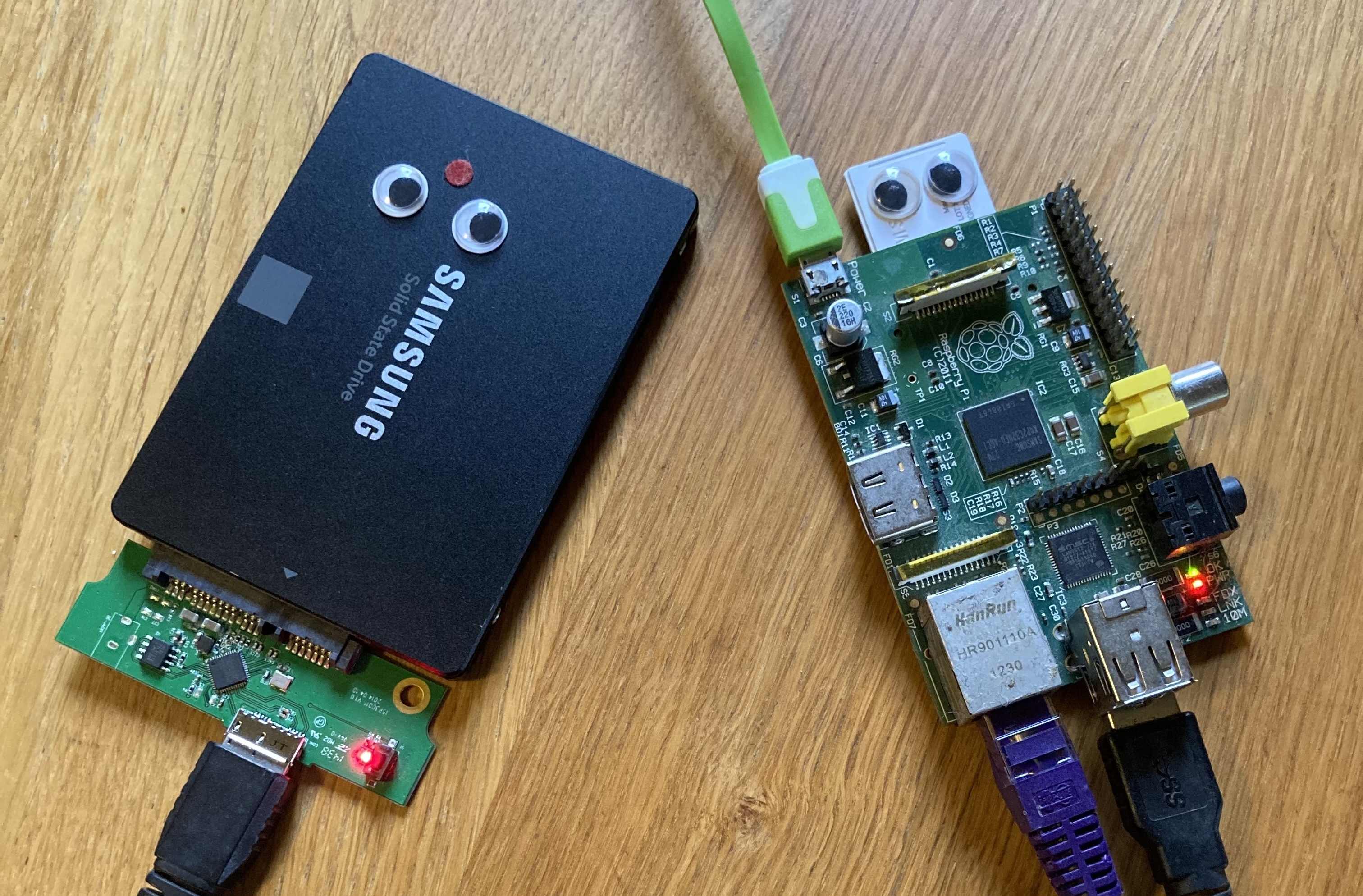 A black 2.5" Samsung SSD with googly eyes lies appears to look at the raspberry pi original model b, who also has googly eyes on its protruding SSD. the pi has a bright green power cable, a purple network cable, and a black USB cable joining it to the SSD.

They lie on a light wooden surface under soft natural light.